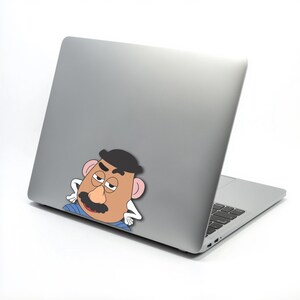 May include: A silver laptop with a Mr. Potato Head sticker on the back. The sticker shows a cartoon character with a tan face, black mustache and hat, and pink ears. The laptop is open, showing the keyboard.