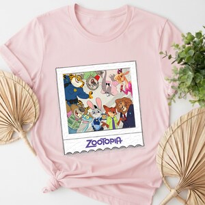 May include: A light pink t-shirt featuring a colorful graphic of characters from the animated movie Zootopia. The graphic is in a polaroid-style frame and includes the movie's title in purple text. The shirt is made of a soft material.