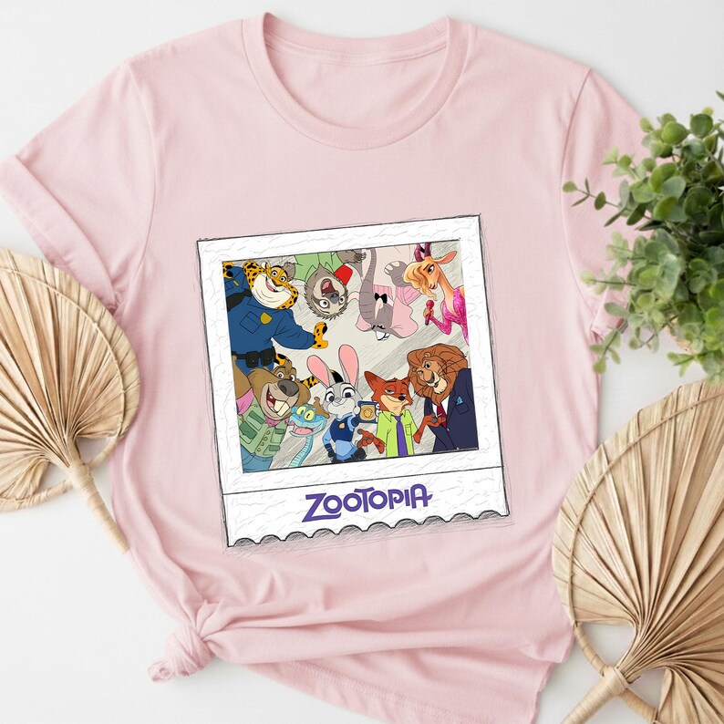 May include: A light pink t-shirt featuring a colorful graphic of characters from the animated movie Zootopia. The graphic is in a polaroid-style frame and includes the movie's title in purple text. The shirt is made of a soft material.