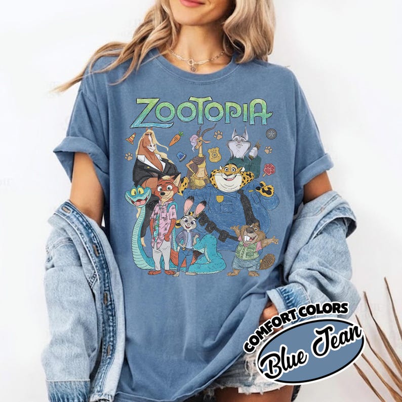 May include: A blue jean colored t-shirt featuring a graphic print of Zootopia characters including Nick Wilde, Judy Hopps, and Chief Bogo. The text Zootopia is printed in green above the characters. The shirt has short sleeves and a crew neckline.
