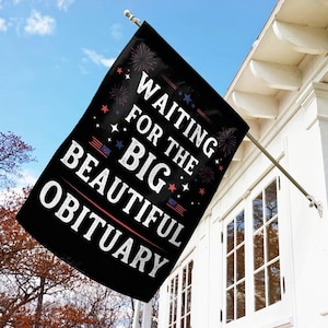 May include: Black flag with white text that reads "WAITING FOR THE BIG BEAUTIFUL OBITUARY." The flag has red, white, and blue accents and is hanging from a silver pole attached to a white building.