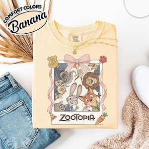 May include: A pale yellow t-shirt featuring a graphic print of a Polaroid-style frame containing cartoon characters from Zootopia, including Judy Hopps and Nick Wilde. The frame is decorated with a pink polka-dot bow, a badge, and a carrot icon.