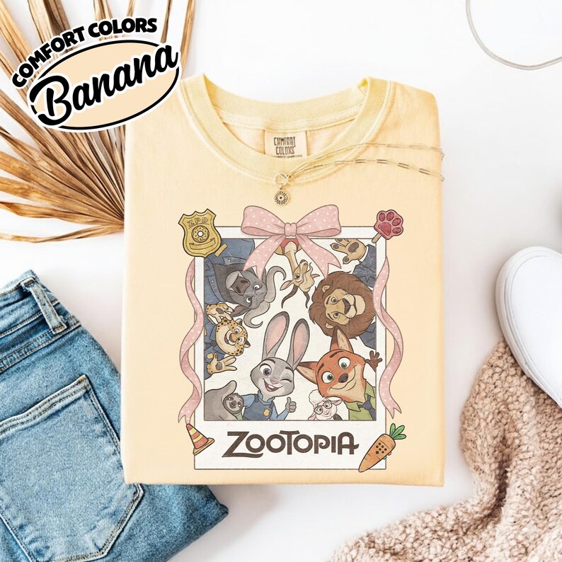 May include: A pale yellow t-shirt featuring a graphic print of a Polaroid-style frame containing cartoon characters from Zootopia, including Judy Hopps and Nick Wilde. The frame is decorated with a pink polka-dot bow, a badge, and a carrot icon.