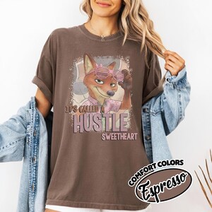 Puede incluir: Una camiseta de manga corta de color marr&oacute;n oscuro con un gr&aacute;fico de un zorro de dibujos animados con gafas de sol rosas y una camisa rosa. El texto dice It's called a hustle sweetheart. La camiseta tiene cuello redondo y corte holgado.