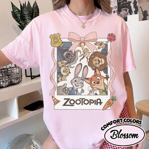 May include: A light pink short-sleeve t-shirt featuring a graphic print of Zootopia characters inside a polaroid-style frame. The design includes a large pink bow at the top, a ZPD badge, a paw print, a traffic cone, and a carrot. The text Zootopia is printed below.