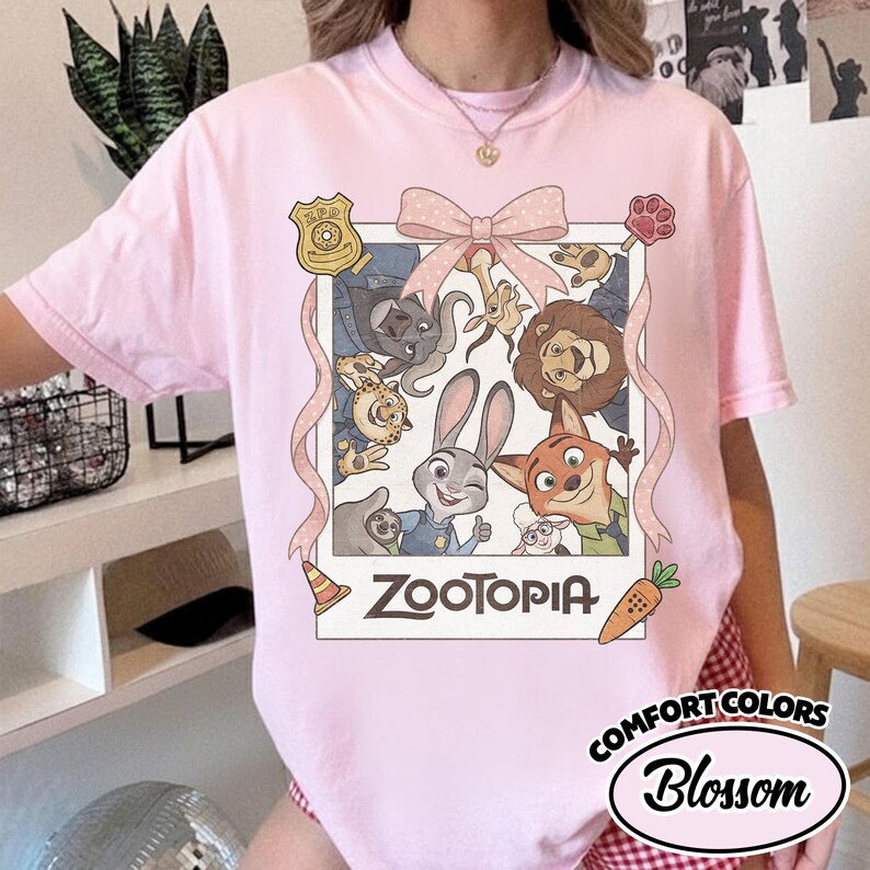 May include: A light pink short-sleeve t-shirt featuring a graphic print of Zootopia characters inside a polaroid-style frame. The design includes a large pink bow at the top, a ZPD badge, a paw print, a traffic cone, and a carrot. The text Zootopia is printed below.