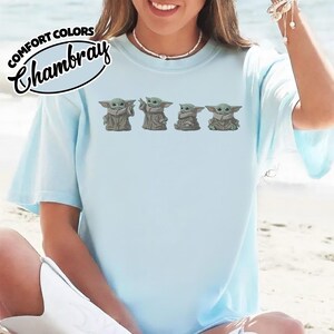 Puede incluir: Una camiseta de cuello redondo azul claro con un estampado gr&aacute;fico de cuatro peque&ntilde;as criaturas verdes con t&uacute;nicas color canela. El texto Comfort Colors Chambray est&aacute; impreso en negro en la esquina superior izquierda. La camiseta tiene mangas cortas.