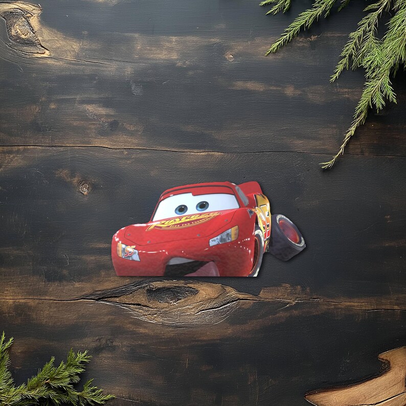 May include: A red Lightning McQueen car-shaped decoration with blue eyes and a smiling mouth. The car has a detached tire and is set against a dark wood background with green sprigs.