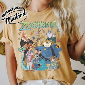 May include: A mustard yellow t-shirt featuring a graphic print of Zootopia characters including Judy Hopps, Nick Wilde, and Chief Bogo. The text "ZOOTOPIA" appears in a stylized font above the characters. A "COMFORT COLORS MUSTARD" label is visible.