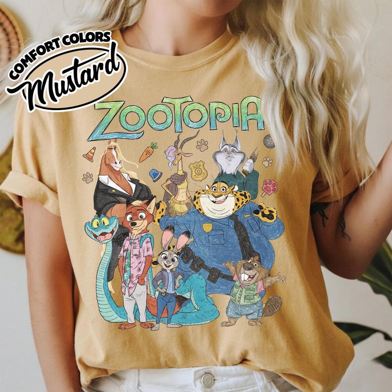 May include: A mustard yellow t-shirt featuring a graphic print of Zootopia characters including Judy Hopps, Nick Wilde, and Chief Bogo. The text "ZOOTOPIA" appears in a stylized font above the characters. A "COMFORT COLORS MUSTARD" label is visible.