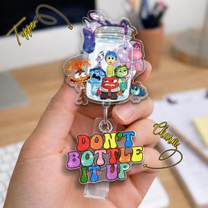 May include: A clear acrylic badge reel featuring characters from the movie "Inside Out" inside a jar. The badge reel has the text "DON'T BOTTLE IT UP" in colorful bubble letters. A silver clip and retractable cord are attached.