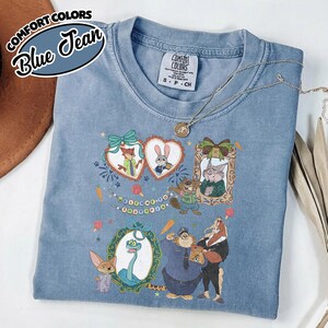 May include: A blue jean colored t-shirt featuring a graphic print of Zootopia characters including Nick Wilde, Judy Hopps, and others in decorative frames. The shirt has a crew neckline and a Comfort Colors brand label on the inside collar area.