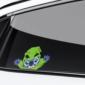 May include: A car window decal featuring a cartoon character, Stitch, dressed as Oogie Boogie. The decal shows Stitch's blue face and large eyes peeking out from a green Oogie Boogie costume, with his arms outstretched. The decal is on a car window.