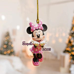 May include: A Minnie Mouse ornament with a pink polka dot bow and dress. The ornament has the name "Sophia" and the year "2025" printed on it. It is hanging from a gold string.