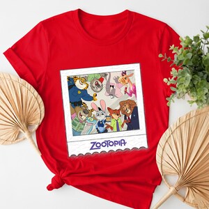 May include: A red t-shirt featuring a graphic of characters from the animated film "Zootopia" in a polaroid-style frame. The frame has a white border and the word "Zootopia" in blue script. The shirt is made of a soft material.