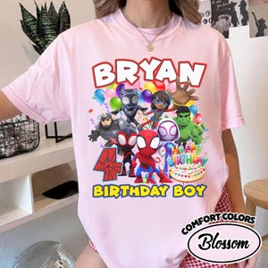 May include: A light pink t-shirt featuring a graphic print of various superhero characters, a birthday cake, and the text "BRYAN", "4", and "BIRTHDAY BOY". The design includes colorful balloons and confetti, with the shirt having a crew neckline.