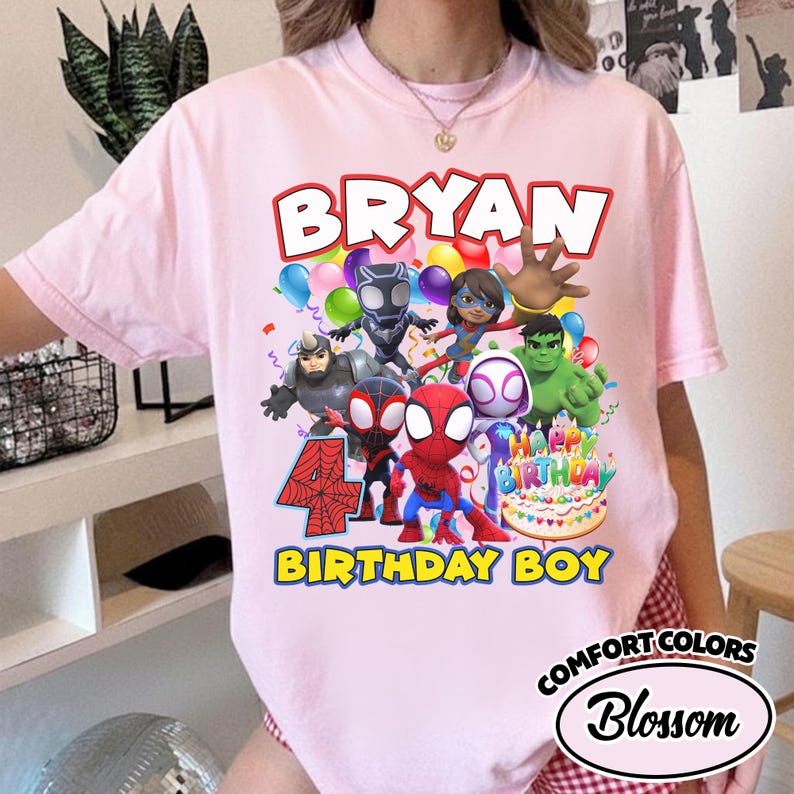 May include: A light pink t-shirt featuring a graphic print of various superhero characters, a birthday cake, and the text "BRYAN", "4", and "BIRTHDAY BOY". The design includes colorful balloons and confetti, with the shirt having a crew neckline.