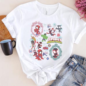 May include: White t-shirt featuring a colorful graphic of Spider-Man and other characters from the "Spidey and His Amazing Friends" animated series. The design includes the show's title and various illustrations of the characters in action poses.