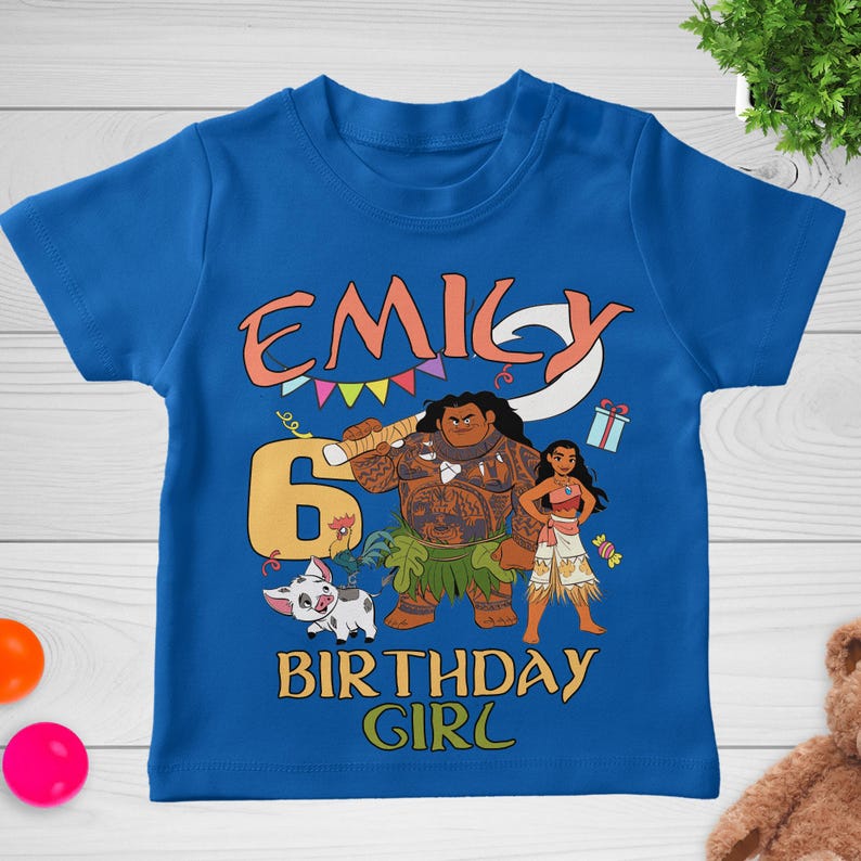 May include: A royal blue short-sleeve t-shirt featuring a graphic print of Moana characters Maui, Moana, and Pua. The design includes the name EMILY, the number 6, and the text BIRTHDAY GIRL in yellow and orange, accented with colorful party decor.