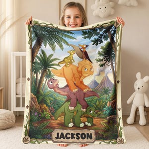 May include: A rectangular fleece blanket featuring a colorful illustration of five cartoon dinosaurs stacked on top of each other in a prehistoric jungle setting with a volcano in the background. The name JACKSON is printed in black on a stone-like plaque.