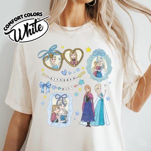 May include: A cream-colored t-shirt featuring a graphic print of characters from Frozen, including Anna and Elsa in frames, a heart-shaped locket, blue bows, snowflakes, and a string of beads with the text birthday girl printed in black letters.