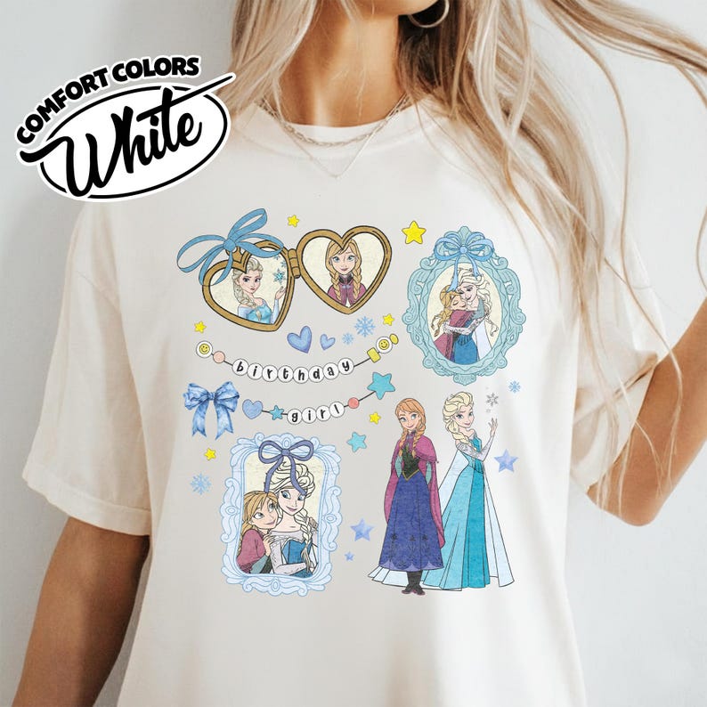 May include: A cream-colored t-shirt featuring a graphic print of characters from Frozen, including Anna and Elsa in frames, a heart-shaped locket, blue bows, snowflakes, and a string of beads with the text birthday girl printed in black letters.