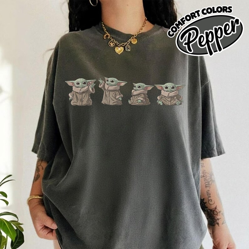 Puede incluir: Una camiseta gris carb&oacute;n con un estampado gr&aacute;fico de cuatro peque&ntilde;as criaturas verdes con t&uacute;nicas color canela dispuestas en una fila horizontal en el pecho. El texto COMFORT COLORS Pepper aparece en una burbuja ovalada blanca en la esquina superior derecha.