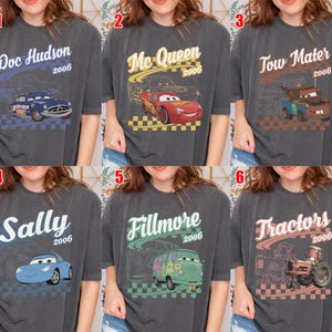 May include: A collection of six charcoal gray t-shirts featuring characters from the 2006 film Cars. Each shirt displays a unique character graphic, including Doc Hudson, McQueen, Tow Mater, Sally, Fillmore, and Tractors, with names and the year 2006.