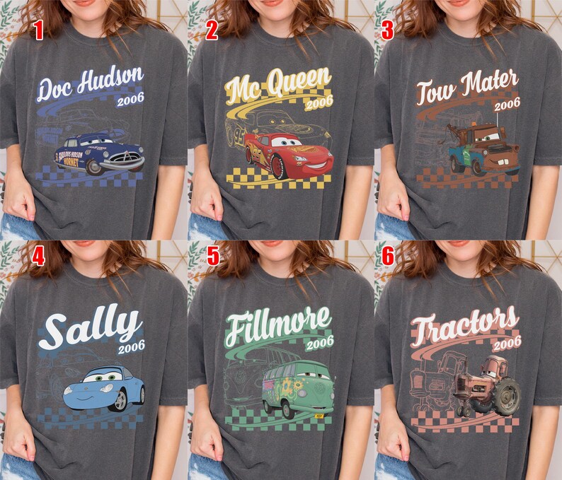 May include: A collection of six charcoal gray t-shirts featuring characters from the 2006 film Cars. Each shirt displays a unique character graphic, including Doc Hudson, McQueen, Tow Mater, Sally, Fillmore, and Tractors, with names and the year 2006.