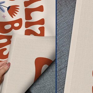 May include: Off-white fabric featuring a retro floral design and the words "California Breeze" in orange. The design includes orange flowers, blue and pink leaves, and a star. The fabric is being held up by a hand.