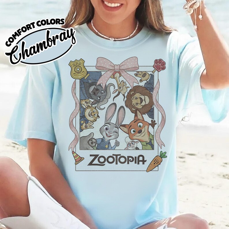 May include: A light blue t-shirt featuring a graphic print of Zootopia characters arranged in a polaroid-style frame with a pink bow at the top. The text Zootopia is printed at the bottom of the frame. The shirt is labeled Comfort Colors Chambray.