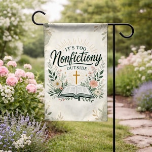May include: A garden flag with the text "IT'S TOO Nonfictiony OUTSIDE" in dark green script. The flag features an open book, a gold cross, and floral accents. The flag is hanging on a black metal stand in a garden setting.