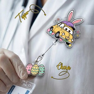 May include: A yellow car-shaped badge reel topper with bunny ears and Easter eggs. The badge reel also has a charm with three decorated Easter eggs. The word "Topper" is written above the car, and the word "Charm" is written below.