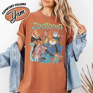 May include: A rust-colored short-sleeve t-shirt featuring a graphic print of Zootopia characters including Judy Hopps, Nick Wilde, and Chief Bogo. The text Zootopia is printed in a stylized font above the characters. The shirt has a crew neckline.