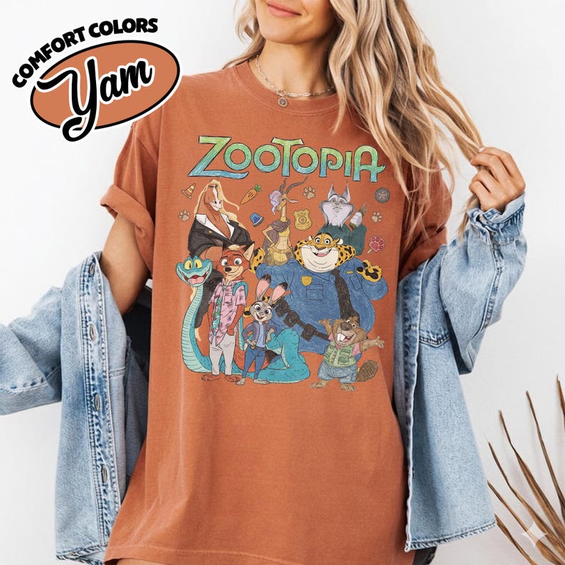 May include: A rust-colored short-sleeve t-shirt featuring a graphic print of Zootopia characters including Judy Hopps, Nick Wilde, and Chief Bogo. The text Zootopia is printed in a stylized font above the characters. The shirt has a crew neckline.
