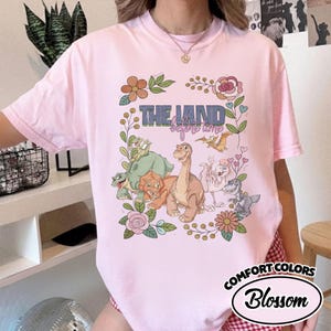 May include: A light pink t-shirt featuring a graphic print of cartoon dinosaurs from The Land Before Time, surrounded by floral illustrations. The text THE LAND before time is printed in blue and pink above the dinosaur characters.
