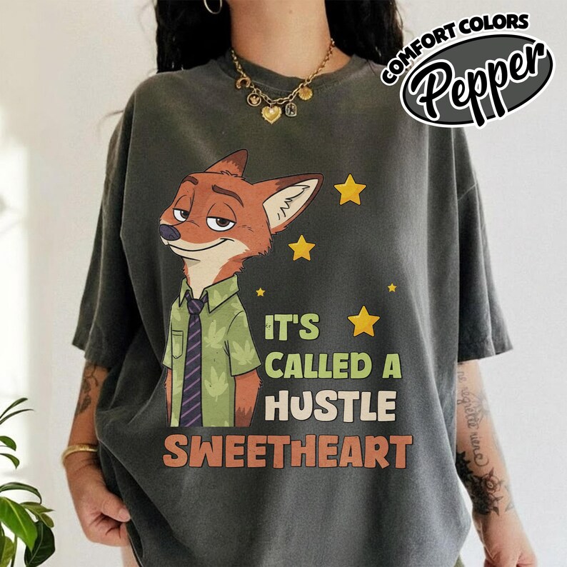 Puede incluir: Una camiseta de manga corta gris carb&oacute;n con el gr&aacute;fico de un zorro de dibujos animados con camisa verde y corbata morada. El texto dice IT'S CALLED A HUSTLE SWEETHEART, acompa&ntilde;ado de estrellas amarillas. La prenda tiene una etiqueta Comfort Colors Pepper.