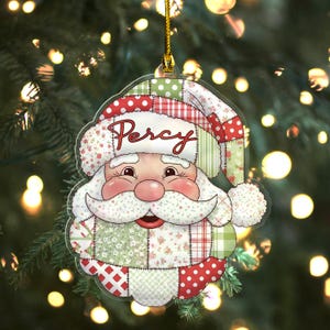 May include: A Christmas ornament shaped like Santa Claus's face. The ornament features a patchwork design in red, green, and white with the name "Percy" in red script. The ornament is hanging from a gold string.