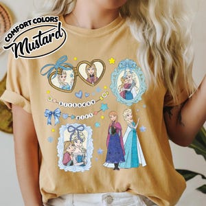 May include: A mustard yellow t-shirt featuring a graphic print of two animated characters in heart-shaped lockets and ornate frames. The design includes blue bows, stars, and a banner with the text "birthday girl" along with the two characters standing.