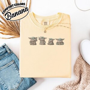 Puede incluir: Una camiseta de cuello redondo de color amarillo p&aacute;lido con un estampado gr&aacute;fico de cuatro peque&ntilde;as criaturas verdes con t&uacute;nicas marrones. La camiseta tiene una etiqueta de la marca Comfort Colors en el interior del cuello.