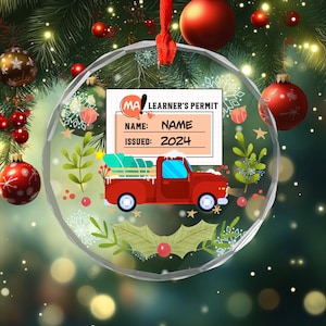 May include: A circular clear acrylic ornament featuring a red pickup truck carrying a Christmas tree. Above the truck is a graphic resembling a learner's permit with the text MA LEARNER'S PERMIT, NAME, and ISSUED: 2024. The design includes holly leaves.