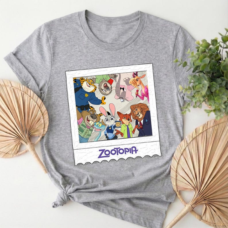 May include: A gray t-shirt featuring a graphic of characters from the animated film "Zootopia" in a polaroid-style frame. The image includes various animal characters in vibrant colors, with the word "Zootopia" printed at the bottom.
