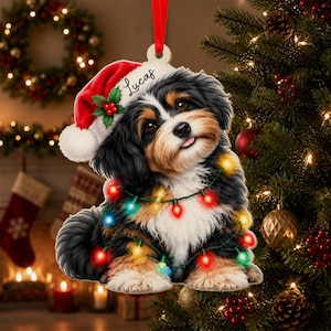 May include: A flat, die-cut Christmas ornament featuring a fluffy black, white, and tan puppy wearing a red Santa hat with a green holly sprig. The puppy is wrapped in colorful string lights and the name Lucas is printed in black script.