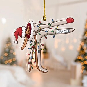 May include: A flat, hanging ornament shaped like a medical speculum, decorated with a red Santa hat with white trim and colorful Christmas string lights. The word NAME is printed in white block letters on the side of the speculum.