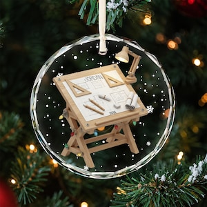 May include: A round, clear glass ornament featuring a wooden drafting table with the name JORDAN and the year 2026 printed on the surface. The table includes a desk lamp, two triangles, pencils, and rolled blueprints, all accented by small colorful lights.