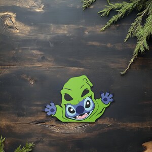May include: A green and blue cartoon character in a green hooded costume with black eye holes and a smiling mouth. The character has blue hands and is set against a dark wood background with green sprigs.