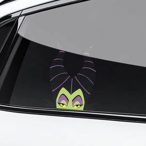 May include: A car window decal featuring a cartoon image of Maleficent, with her signature black horns with purple stripes, green face, and yellow eyes. The decal is visible through the car window.