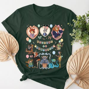 K&ouml;nnte beinhalten: Dunkelgr&uuml;nes T-Shirt mit einer farbenfrohen Grafik aus dem Animationsfilm Zootopia. Das Design umfasst verschiedene Charaktere in herzf&ouml;rmigen Rahmen, Luftballons und den Text "WELCOME TO ZOOTOPIA".