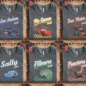 May include: A collection of six dark gray t-shirts featuring characters from the movie Cars. Each shirt displays a unique character name, the year 2006, a graphic of the character, and a checkered pattern at the bottom. Characters include Doc Hudson, Mc Queen, Tow Mater, Sally, Fillmore, and Tractors.
