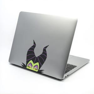 May include: A silver laptop with a black keyboard and a sticker featuring a cartoon character with black horns, green face, and yellow eyes. The sticker is on the back of the laptop.
